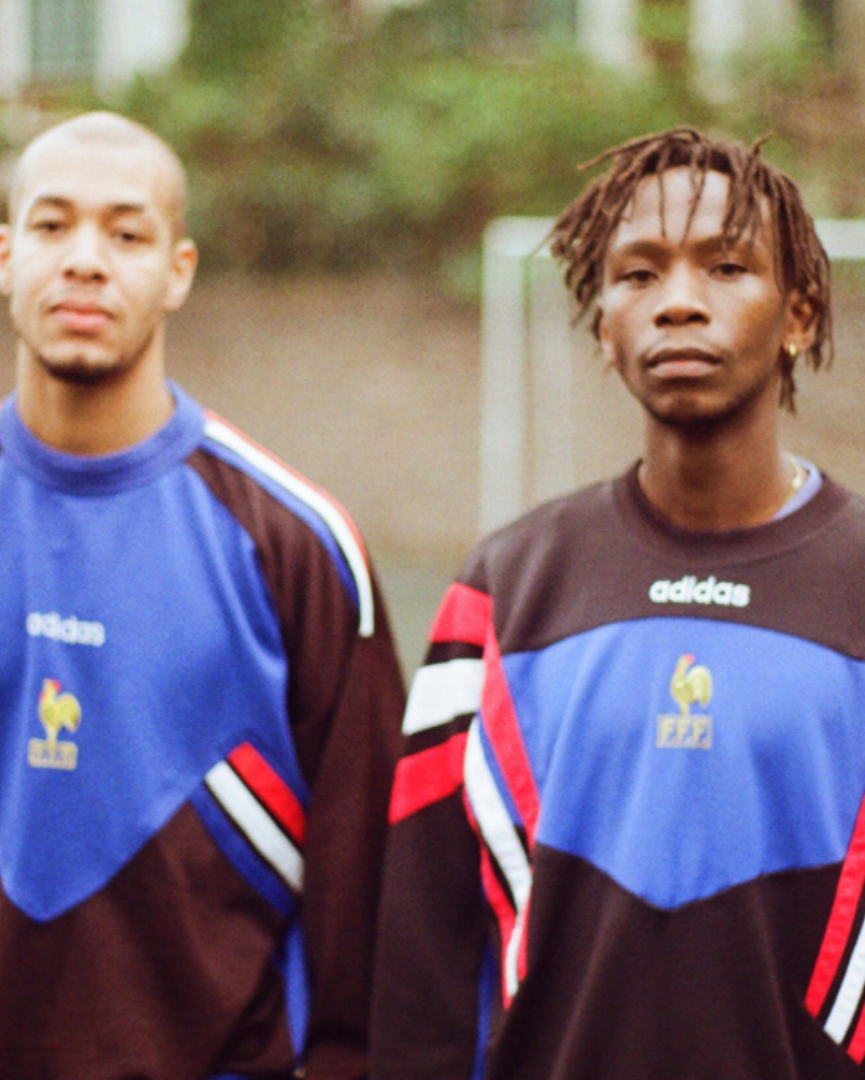 Two men standing on an outdoor sports field wearing matching France national team Adidas long-sleeve athletic shirts with blue, brown, red, and white color blocking.